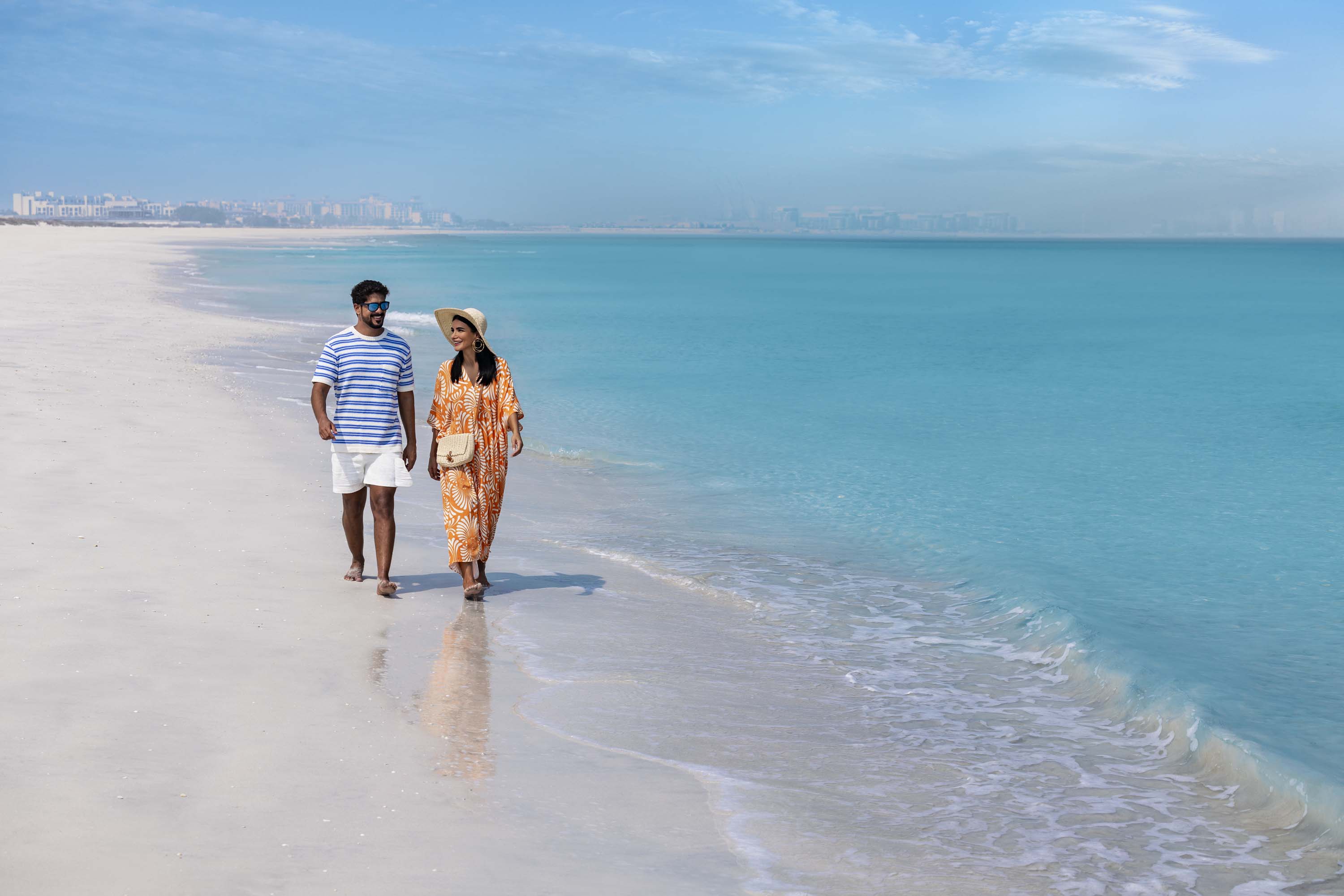 Couple walking barefoot along a serene beach with turquoise water in Abu Dhabi.