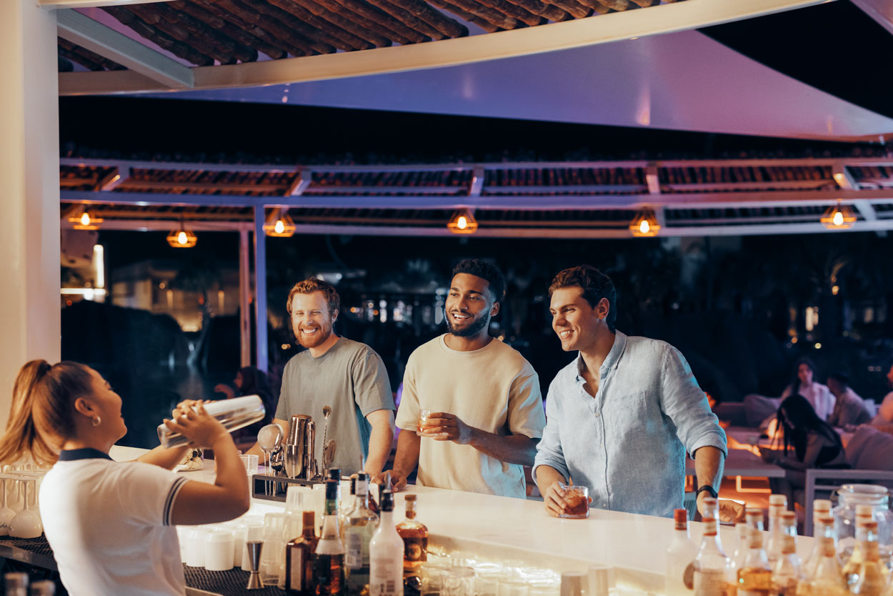Friends enjoying drinks at a lively outdoor bar with a bartender mixing cocktails.