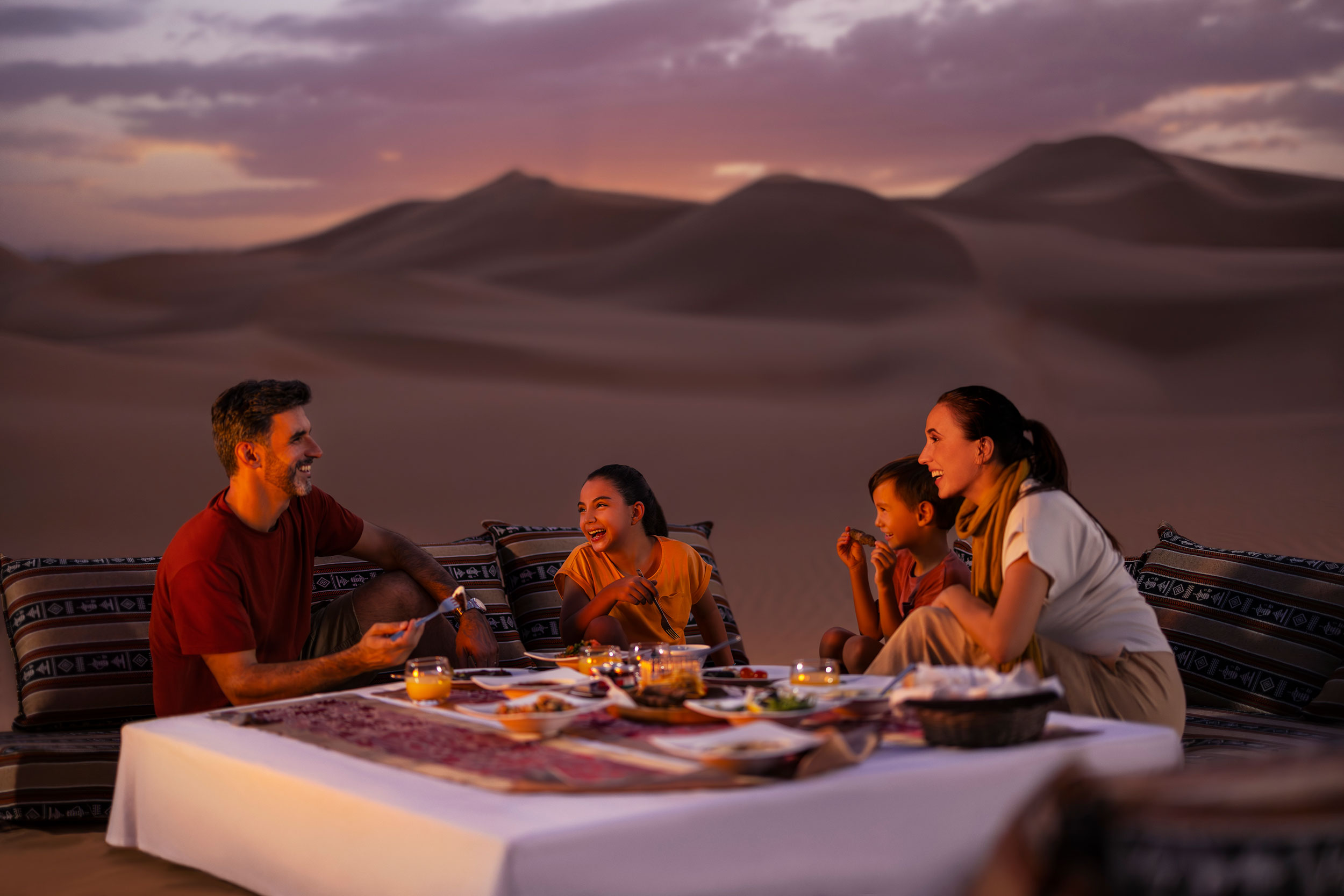 Family enjoying a traditional dinner on cushions in Abu Dhabi desert at sunset with scenic dunes
