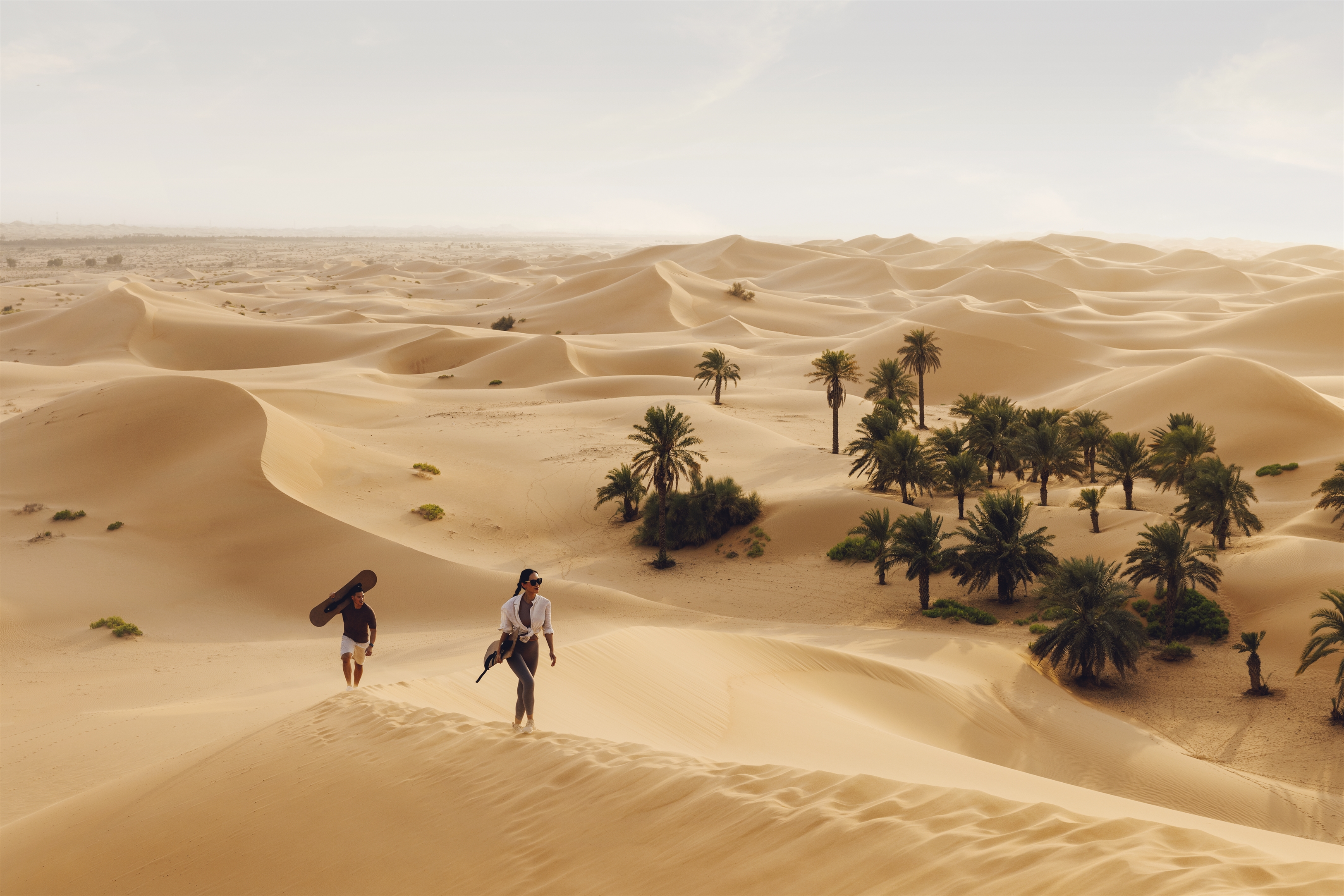 Two people trekking through golden sand dunes near a desert oasis in Abu Dhabi.